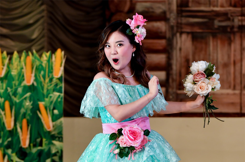 Female opera singer, dressed in a light blue, frilly dress, holding a bouquet of flowers, performing on stage.